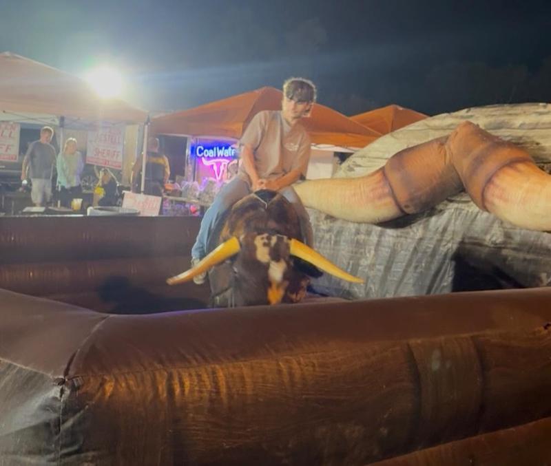An adult rides the Mechanical Bull of the South bull inside the orange inflatable at a night event, with vendor tents and lights behind